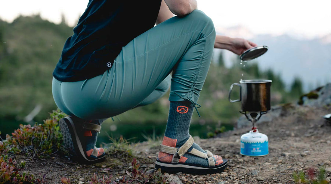 Backpacker wearing Cloudline hiking socks crouched over stove lifting lid of pot.