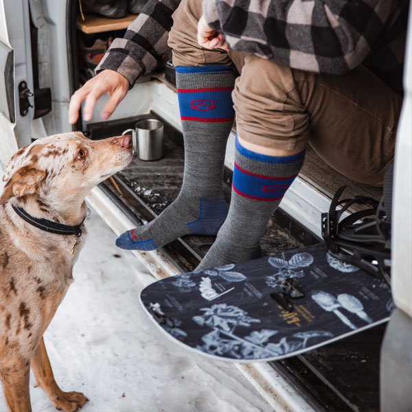 Man wearing Cloudline socks sitting in van sliding doorway next to snowboard and petting dog. 