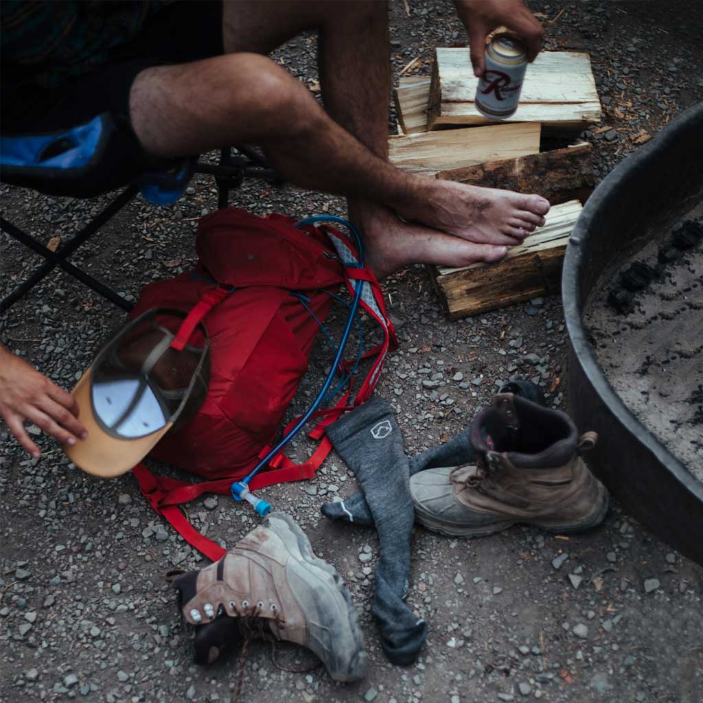 Man with Cloudline compression socks and boots kicked off,  sitting next to campfire with a post hike beer.