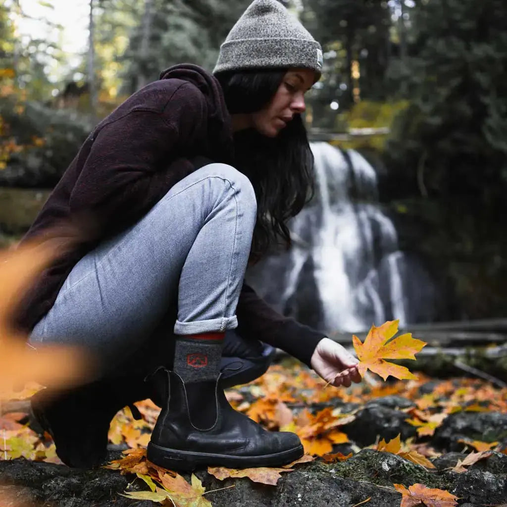 Hiker wearing Cloudline merino wool hiking socks on one knee picking up fall leaves with waterfall in background.