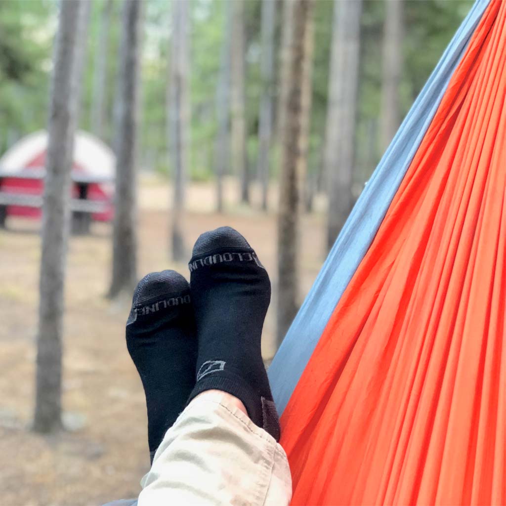 Camper wearing Cloudline running socks laying in hammock with tent and trees in background.