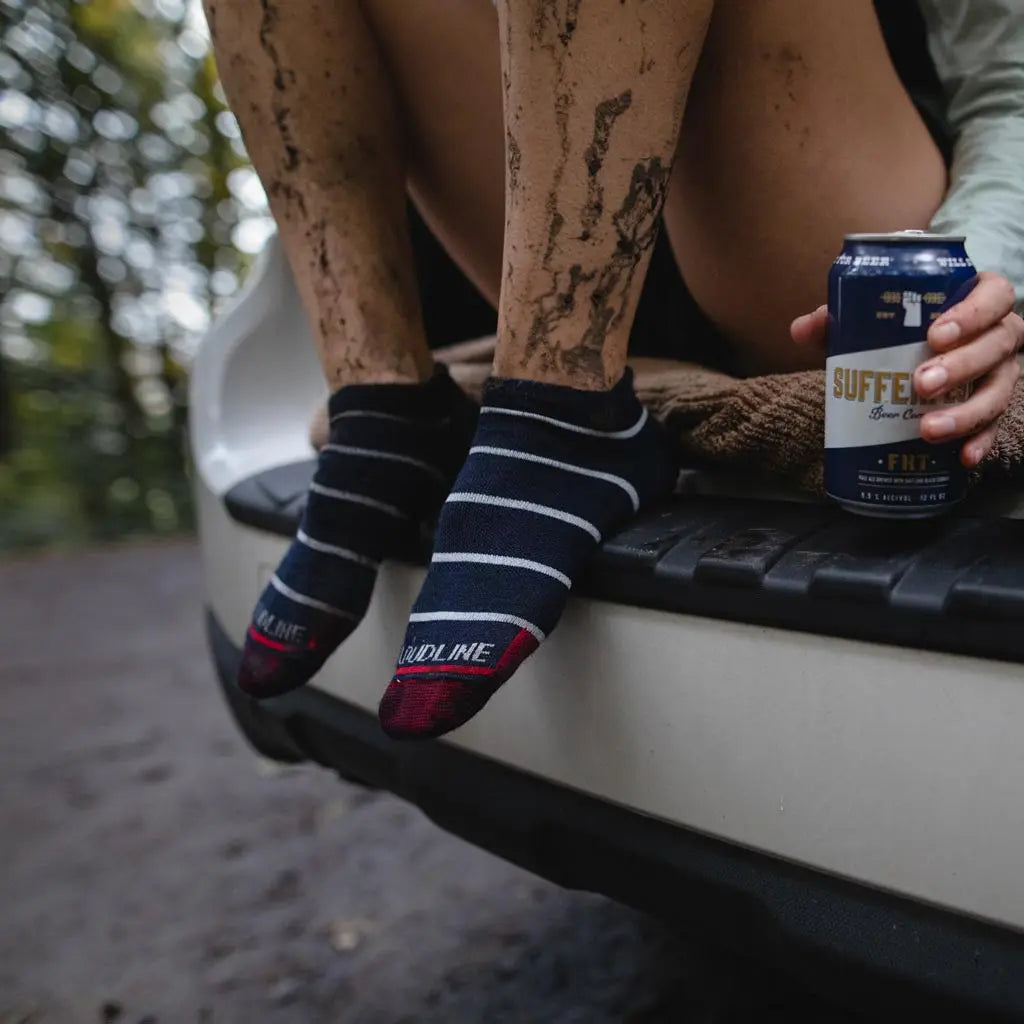 Women wearing Cloudline merino wool running socks sitting in hatchback enjoying a post muddy run beverage with knees up and feet on bumper.