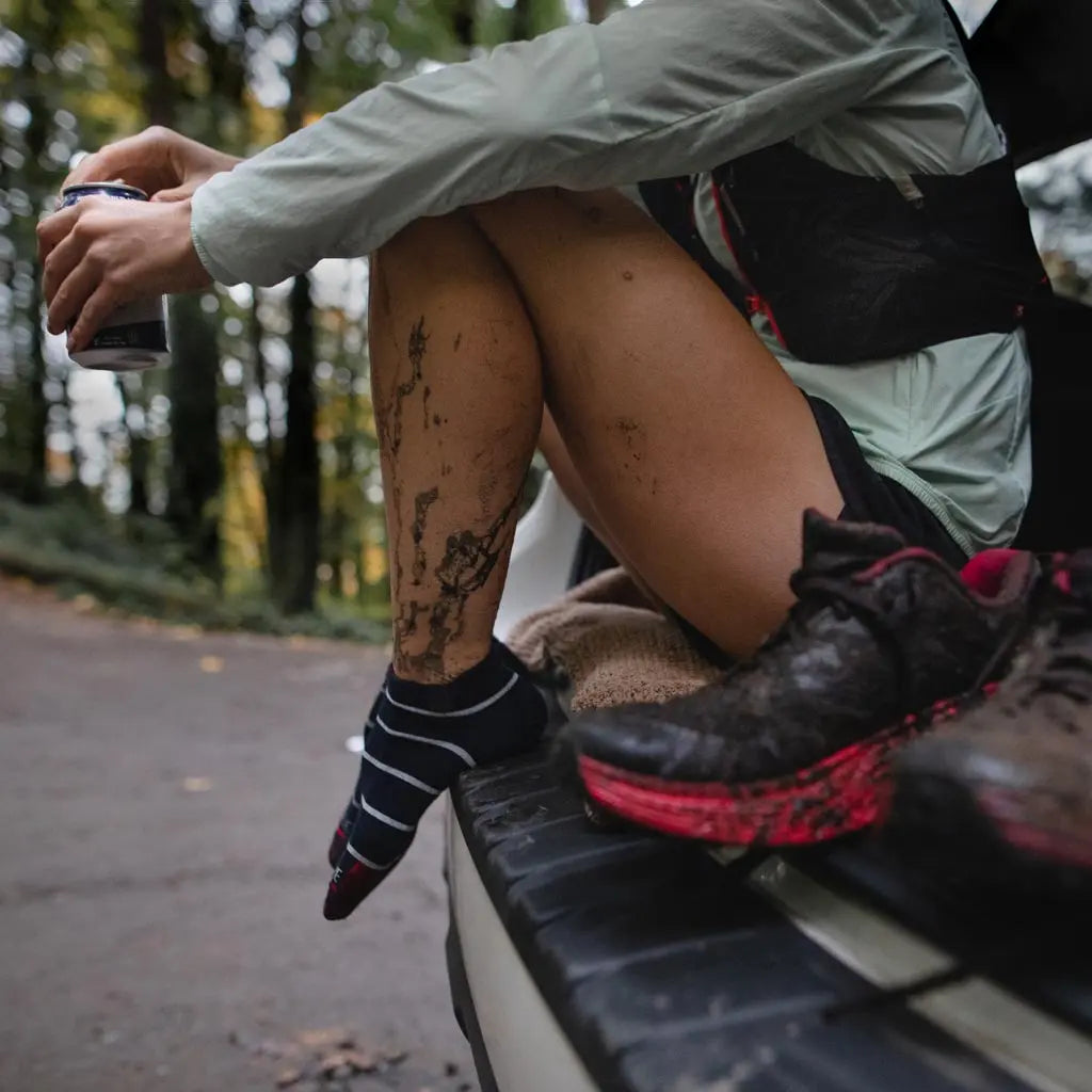 Women wearing Cloudline merino wool no show socks resting on bumper of car after muddy trail run.