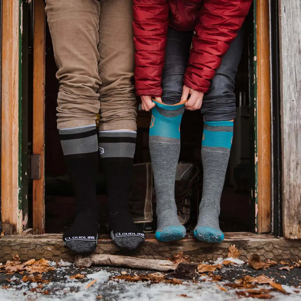 Couple standing in snowy cabin doorway wearing Cloudline merino wool snow socks without shoes.
