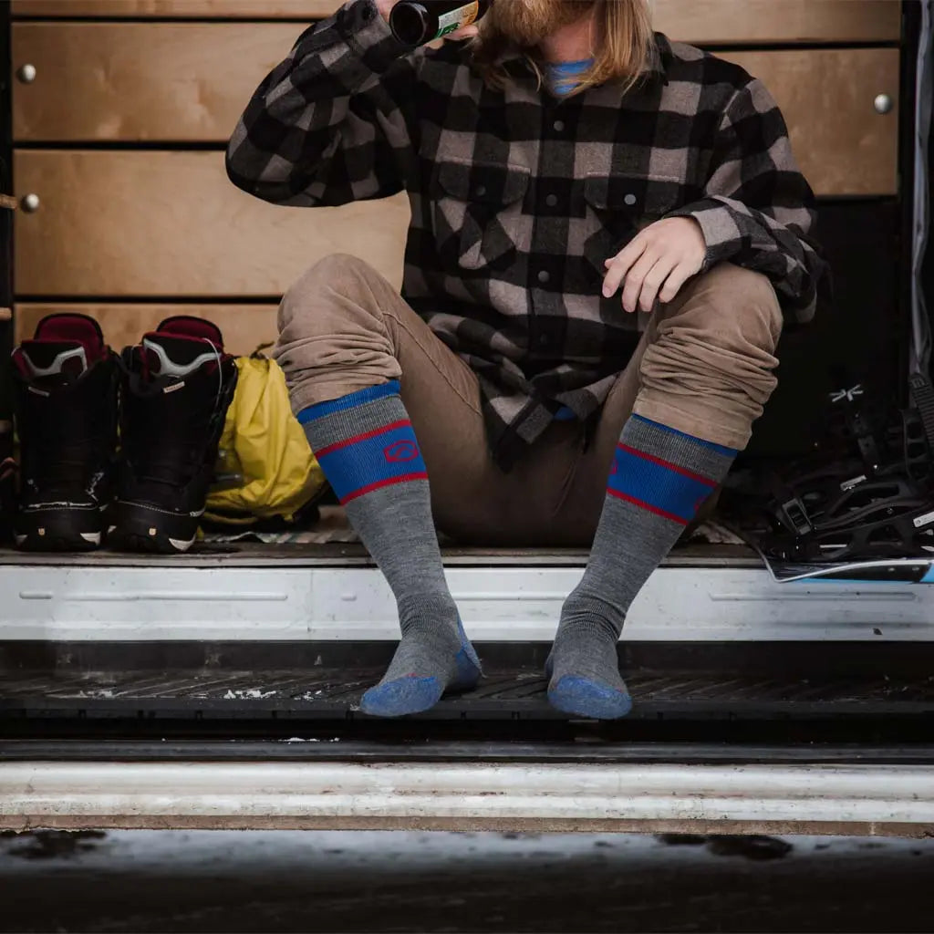 Man wearing Cloudline merino wool snow socks sitting in camper van doorway surrounded by snowboard gear sipping a beverage after a day on the slopes.