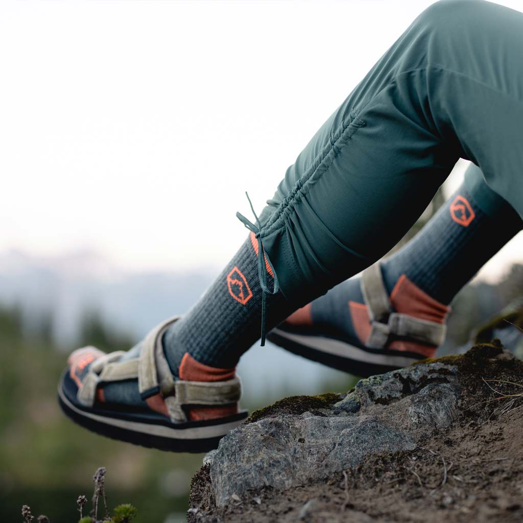 Hiker wearing Cloudline hiking socks and sandals sitting on rock above lake. 