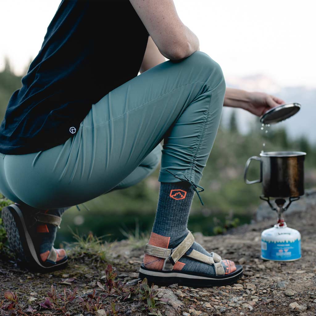 Women wearing Cloudline hiking socks crouched next to backpacking stove lifting pot lid.