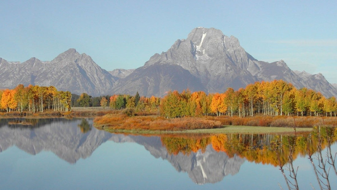Image of a fall sunny day with mountains in the background and a fall-colored forest along a calm lake.