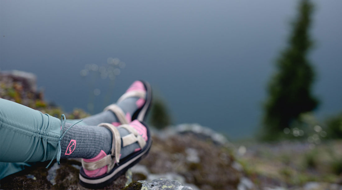 Close up of a hikers feet with Cloudline Apparel socks and sandals with a mountain lake in the background.