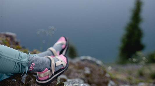 Close up of a hikers feet with Cloudline Apparel socks and sandals with a mountain lake in the background.