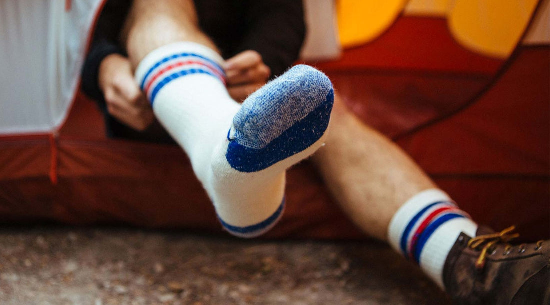 Hiker sitting at a tent pulling on a merino wool crew sock, showing the cushioned sole