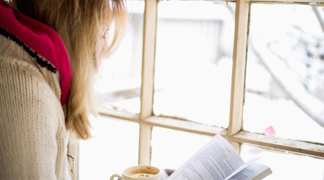 Women in cozy sweater, drinking cocoa and reading by window with winter snow outside.