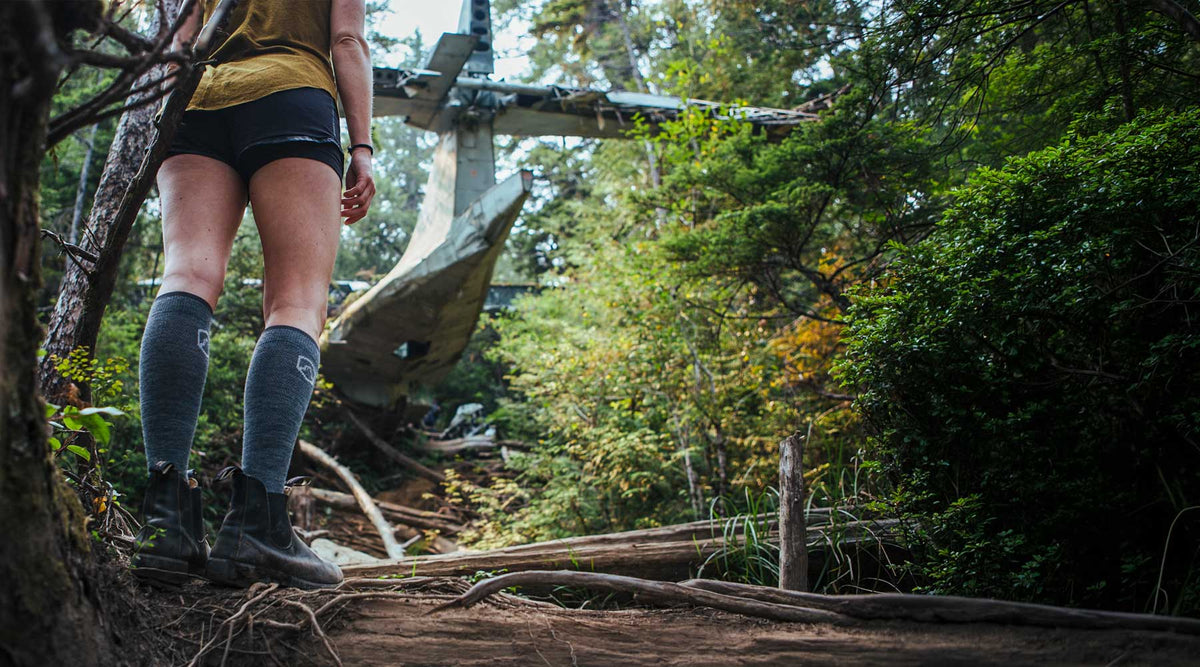 A person hiking on a forest trail, shown from behind wearing athletic shorts, knee-high socks, and hiking boots, with trees and fallen logs in the background.