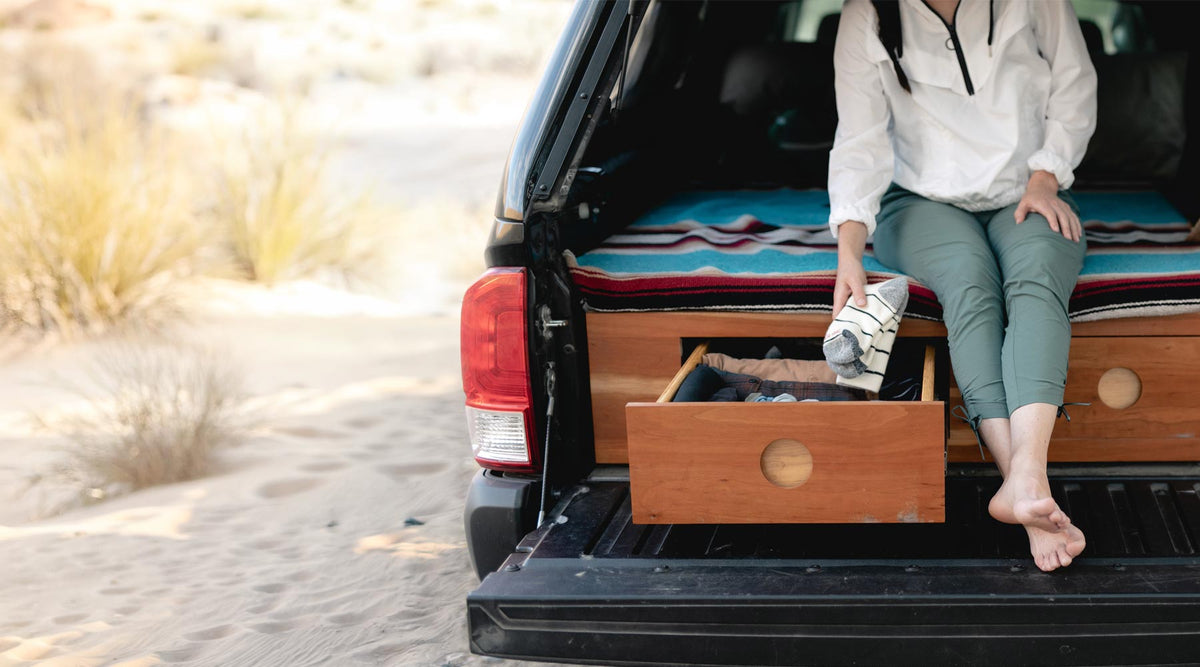 A woman sits barefoot in the open tailgate of a black vehicle, next to a wooden storage drawer with a striped mattress or bed surface behind her, in a sandy outdoor setting.