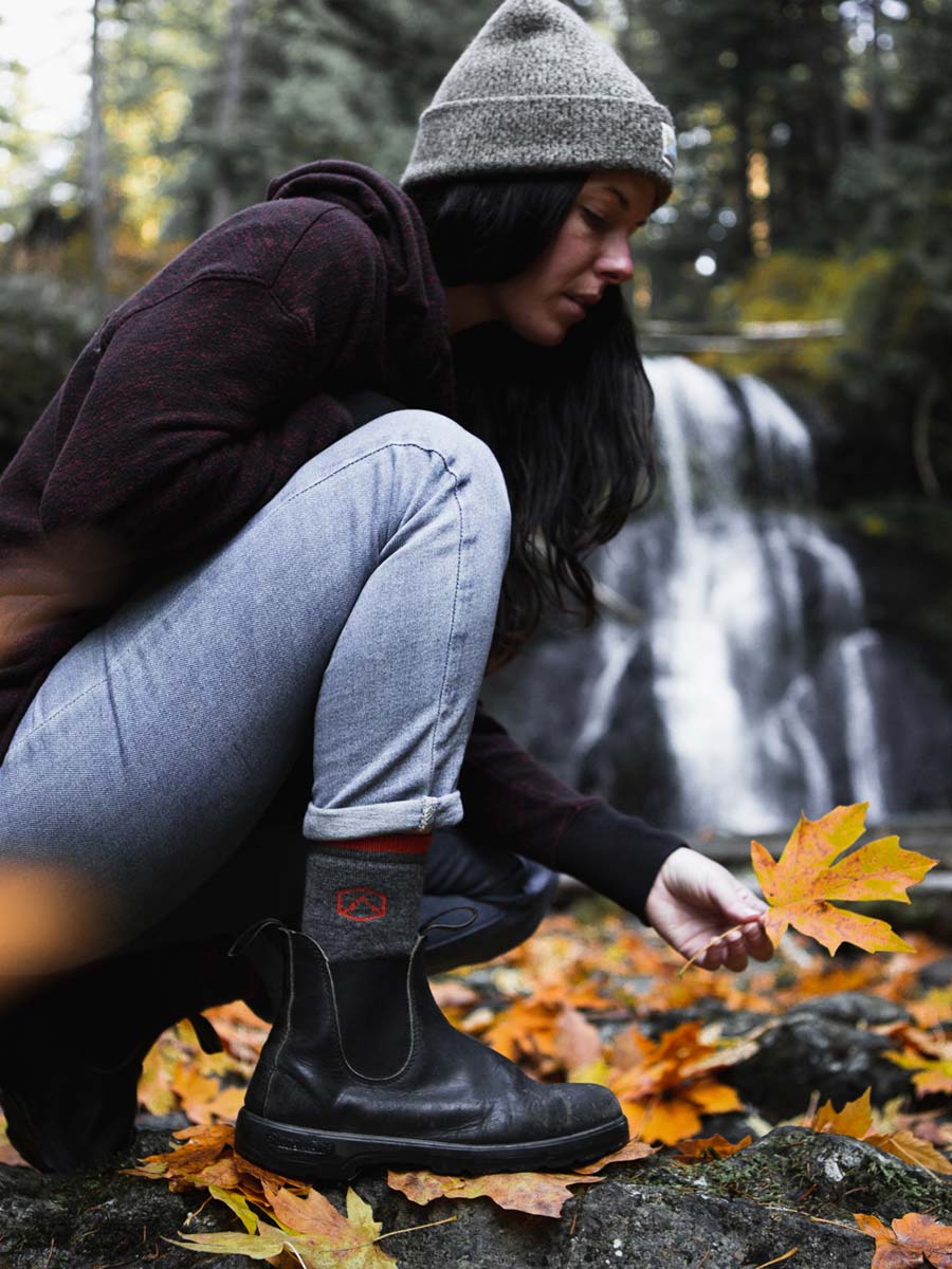 Women wearing Cloudline socks crouching to pick up fall leaf with waterfall in the background. 