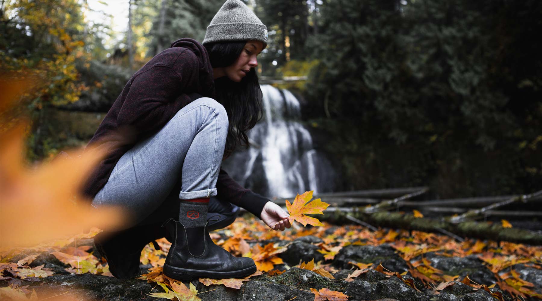 Women wearing Cloudline socks crouching to pick up fall leaf with waterfall in the background. 