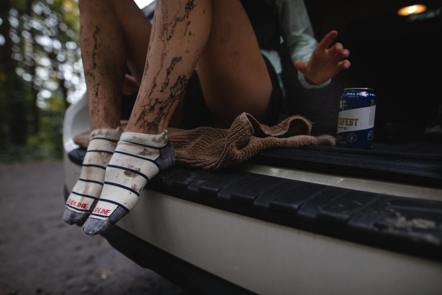 Muddy trail runner wearing Cloudline socks sitting on bumper to take of shoes after run. 