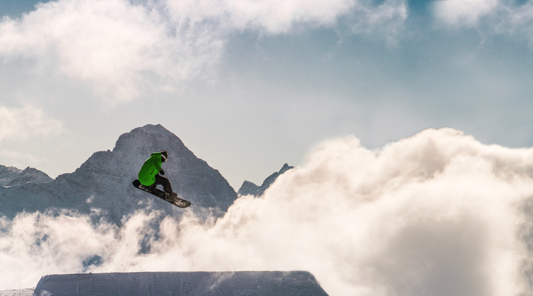 Snowboarder wearing Cloudline socks hitting a large jump with mountains and clouds in the background.