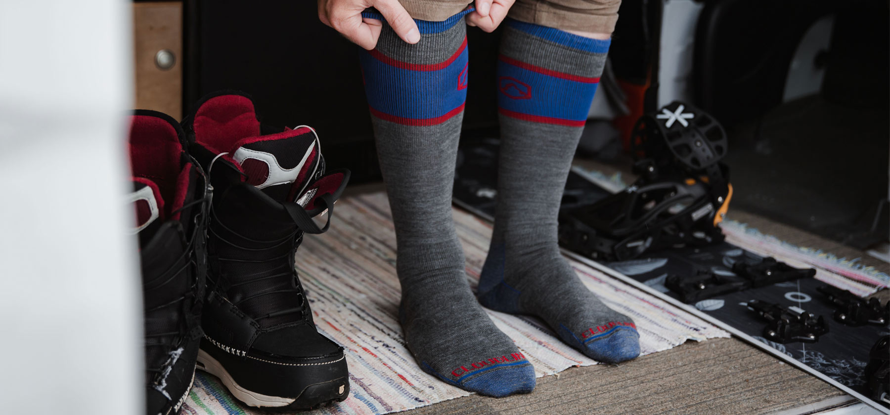 Snowboarder pulling on Cloudline snowboard/ski socks standing inside camper van surrounded by snowboard gear. 