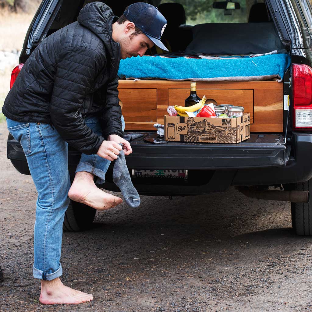 Barefoot man putting on Cloudline hiking socks in front of camper truck. 