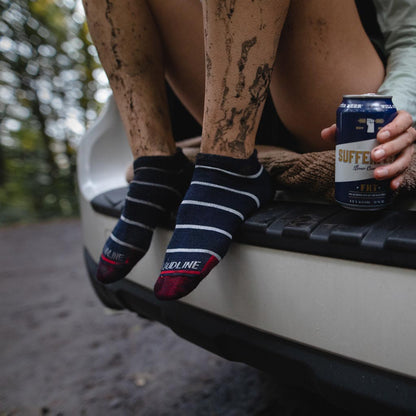 Women wearing Cloudline socks sitting in hatchback enjoying post muddy run beverage with knees up and feet on bumper. 