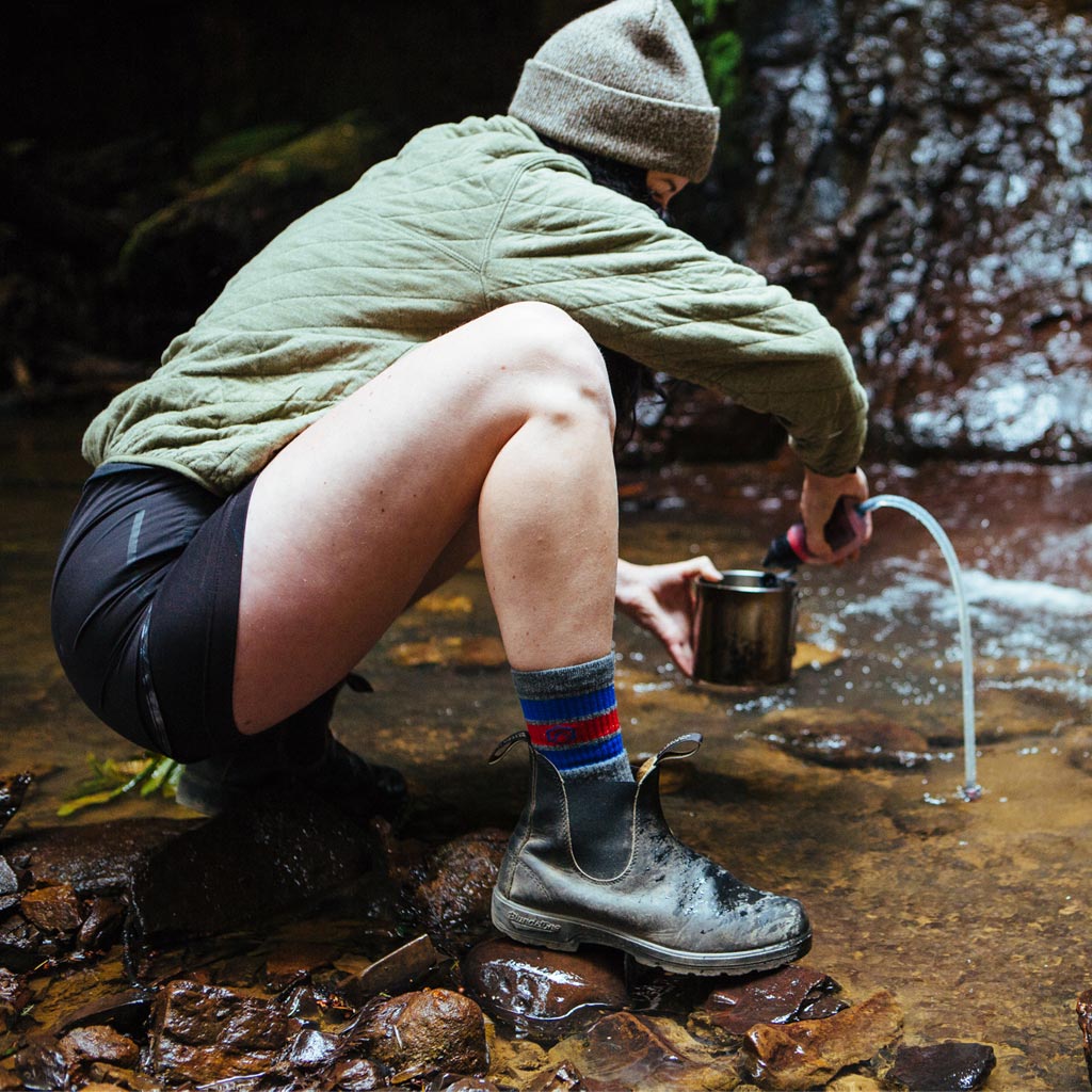 Women wearing Cloudline hiking socks crouched next to stream to filter water into cooking pot.