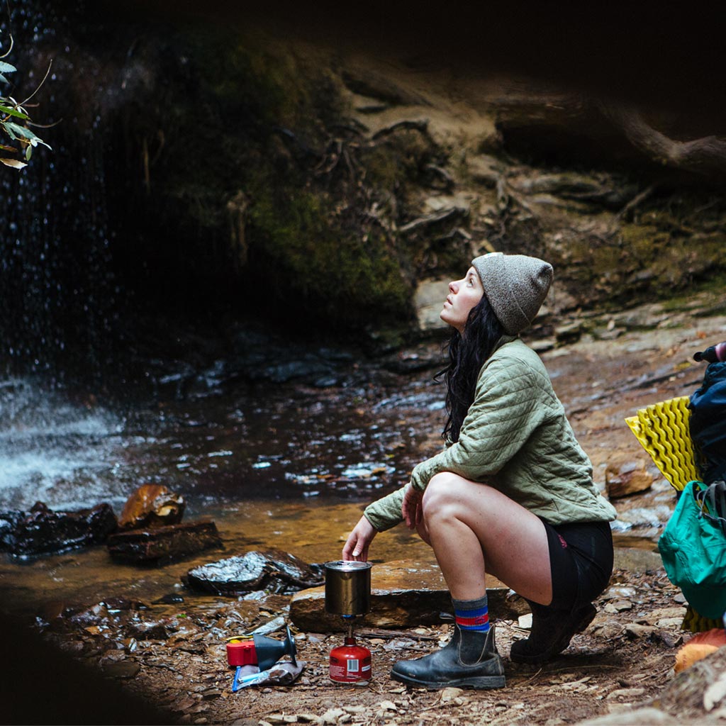 Women wearing Cloudline hiking socks crouched over backpacking stove while dinner cooks. 