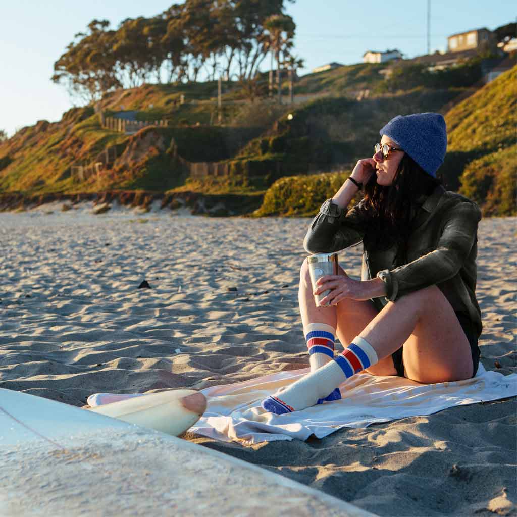 Women wearing Cloudline hiking socks sitting on beach towel next to surf boards.