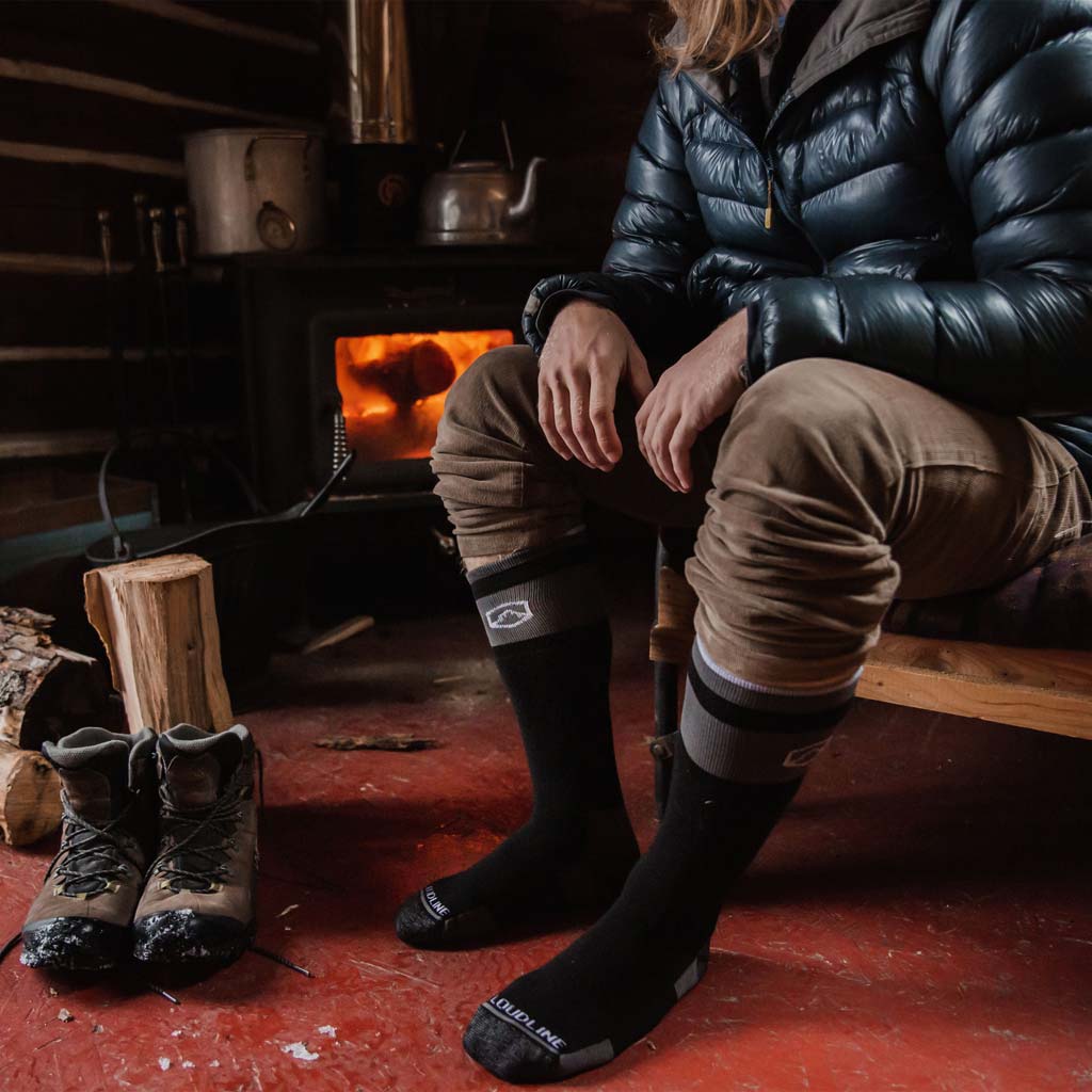 Man sitting in cabin next to wood stove wearing Cloudline snow socks. 