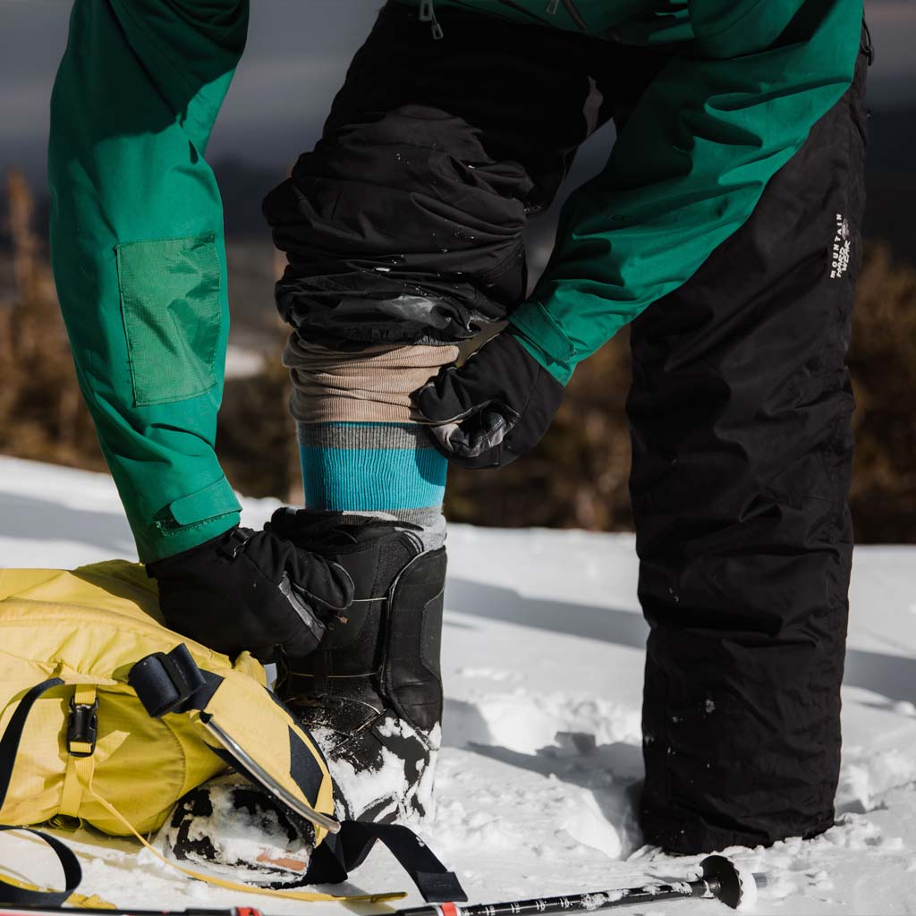 Snowboarder wearing Cloudline socks bending over in the snow to adjust boot. 