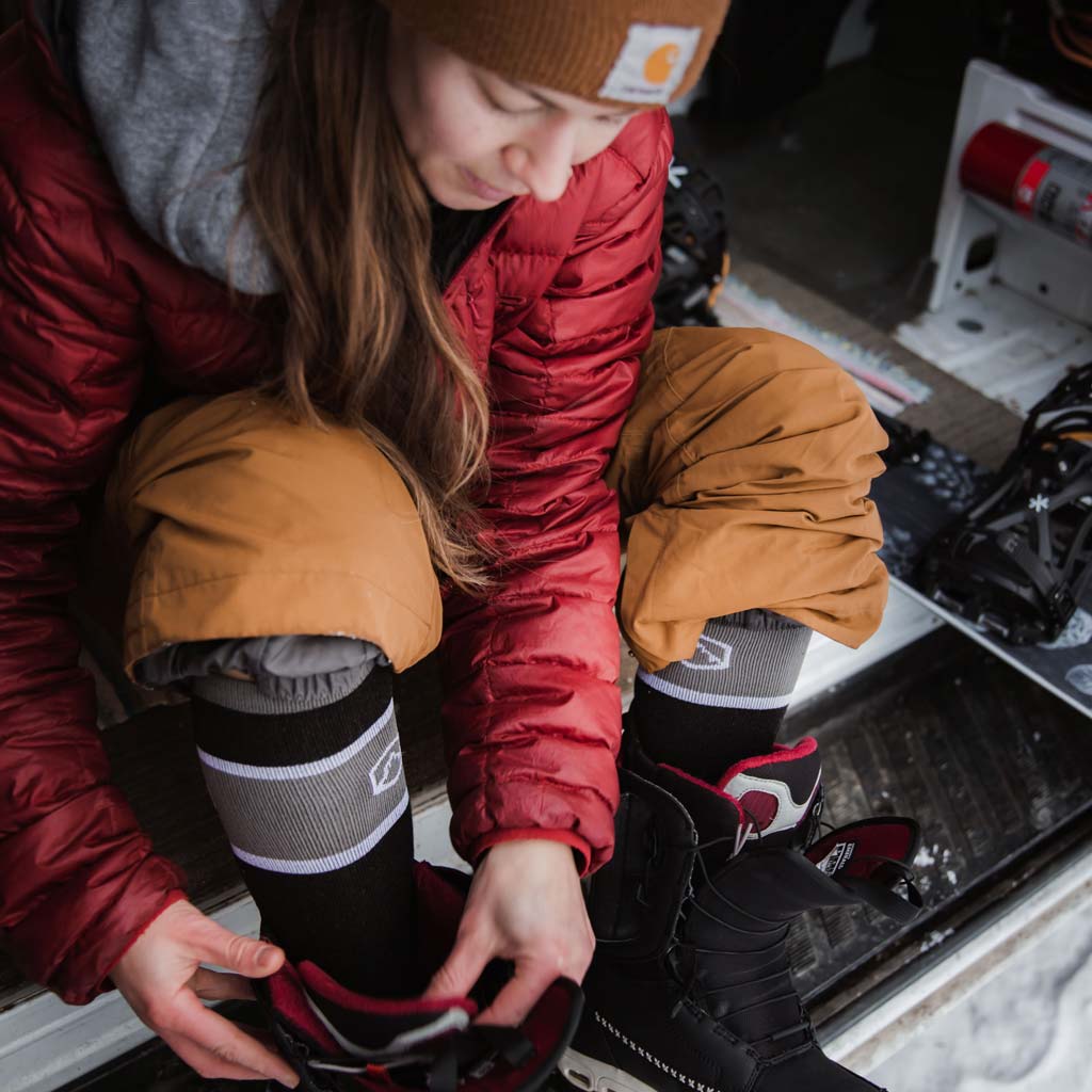 Women wearing Cloudline snow socks putting on boots with snowboard sitting to the side.