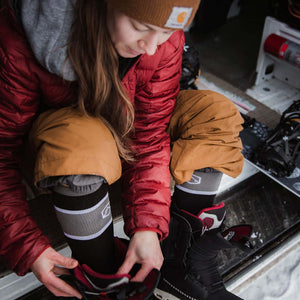 Women wearing Cloudline snow socks putting on boots with snowboard sitting to the side.