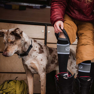 Women wearing Cloudline socks standing next to dog while stepping into snowboard boots.