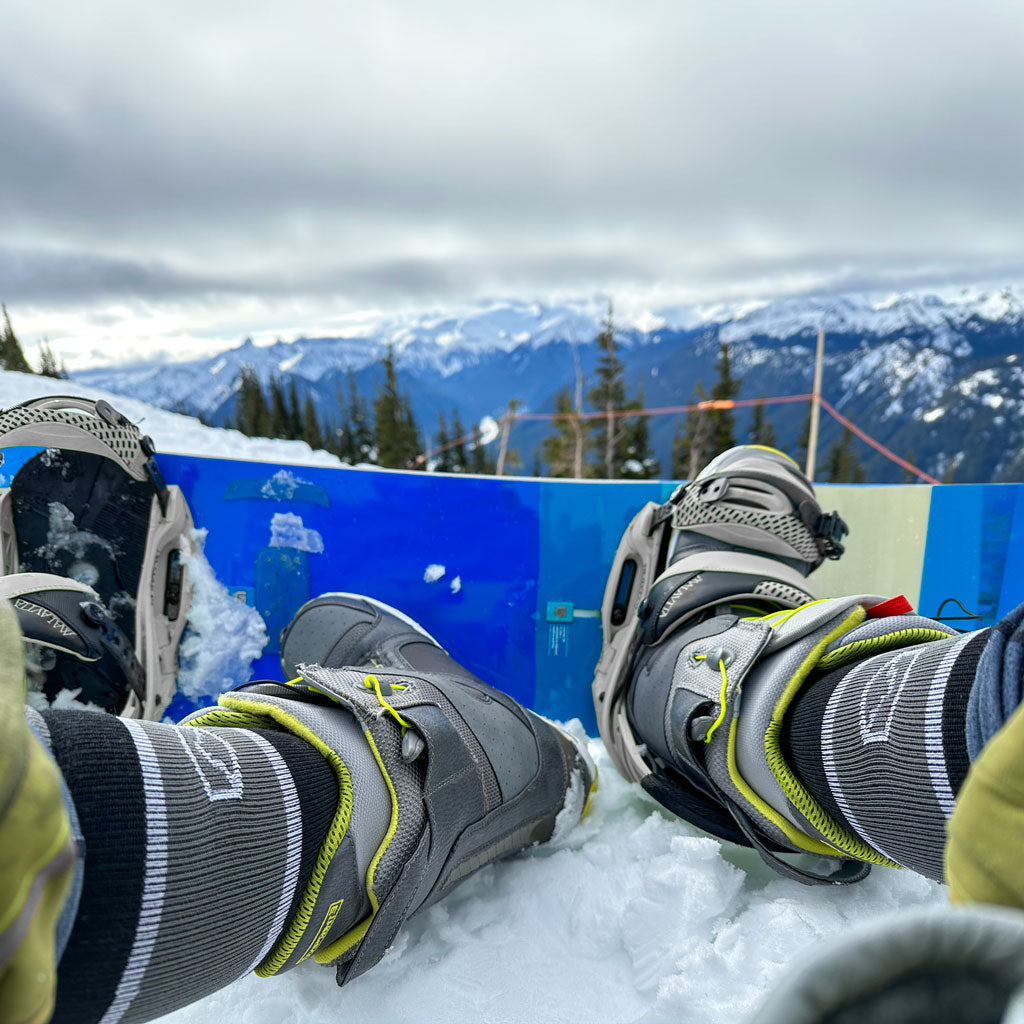 Snowboarder wearing Cloudline socks with one foot out sitting on snow.