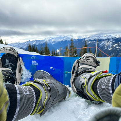 Snowboarder wearing Cloudline socks with one foot out sitting on snow.