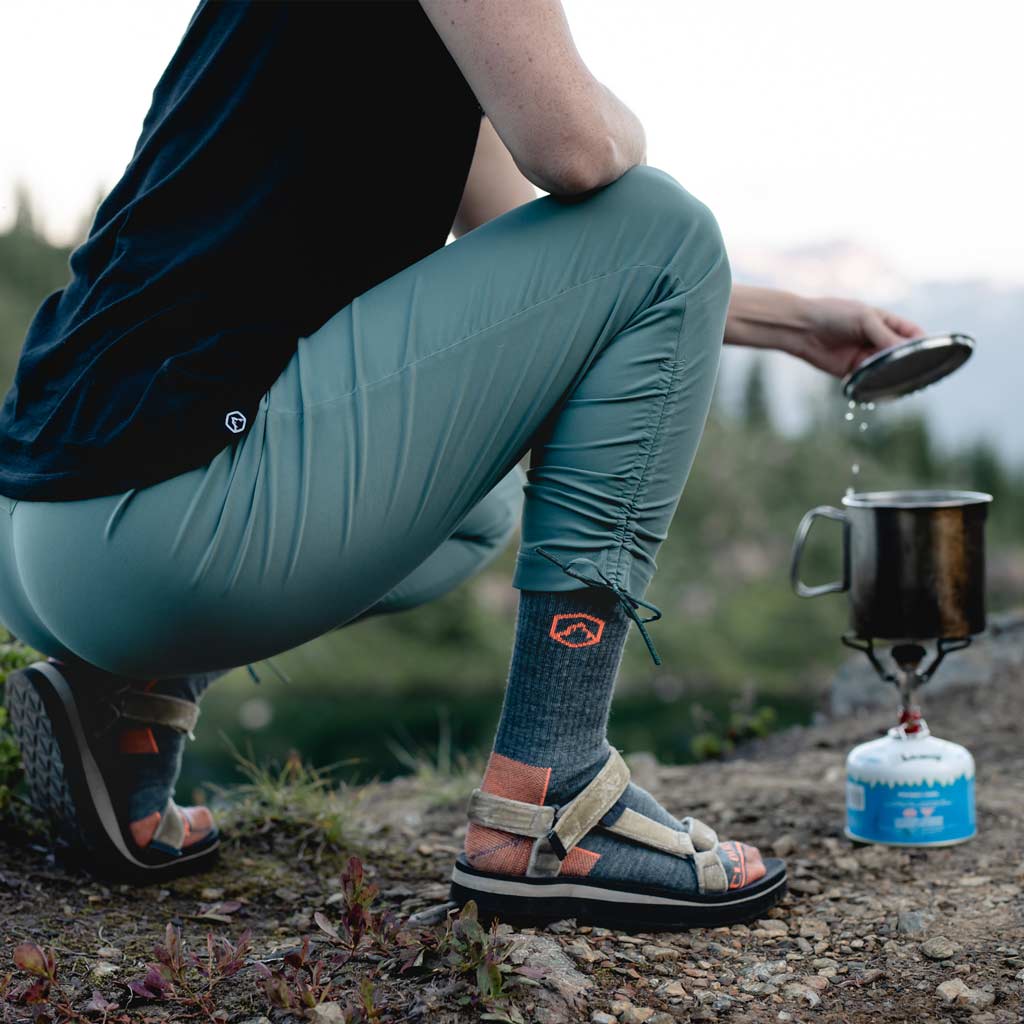 Women wearing Cloudline hiking socks crouched next to backpacking stove lifting pot lid. 
